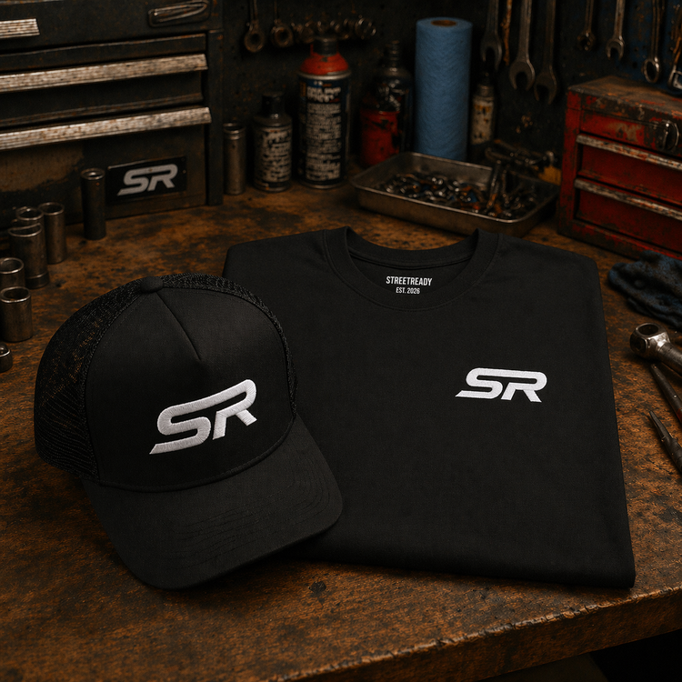 Black StreetReady (SR) logo trucker hat and matching folded black T-shirt displayed on a worn mechanic’s workbench, surrounded by tools, bolts, and shop supplies in a dim garage setting.